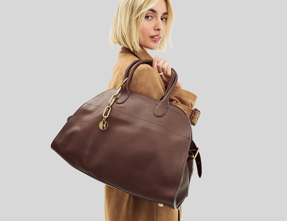A woman with short blonde hair carries a large brown leather duffel bag with gold-tone hardware against a light gray studio background.