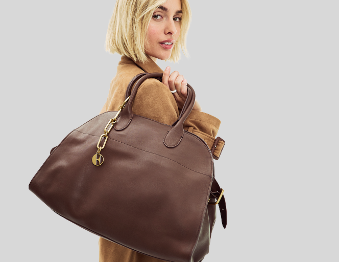 A woman with short blonde hair carries a large brown leather duffel bag with gold-tone hardware against a light gray studio background.