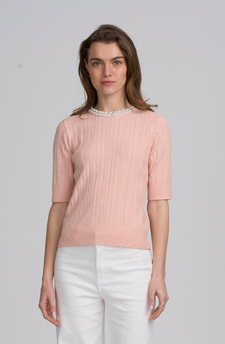 A woman wearing a light pink ribbed knit short-sleeve top with a pearl collar, paired with white pants, photographed against a solid light gray background.