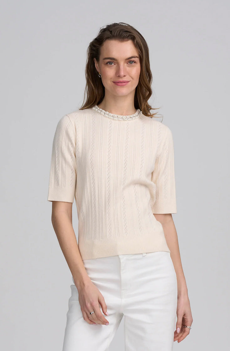A woman modeling a short-sleeve cream-colored knit top with a textured vertical rib pattern and a delicate pearl collar, paired with white pants, against a solid light gray background.