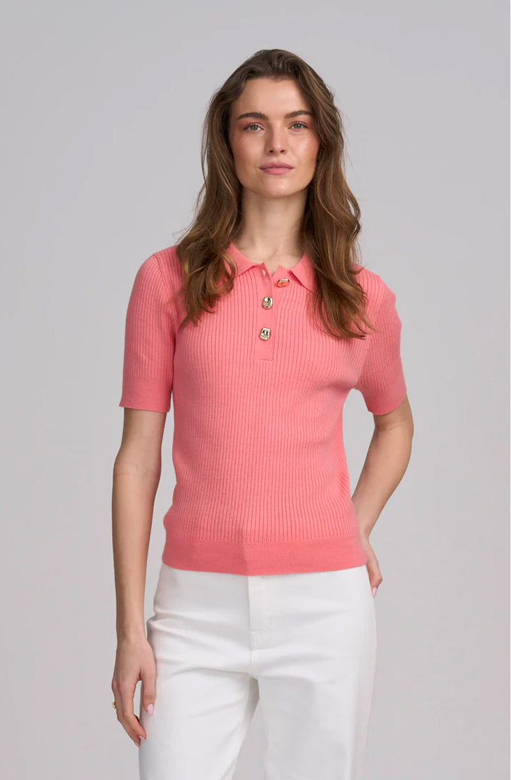 A woman modeling a short-sleeve ribbed knit polo shirt in a coral pink color, featuring gold buttons on the collar, paired with white trousers, against a solid light gray background.