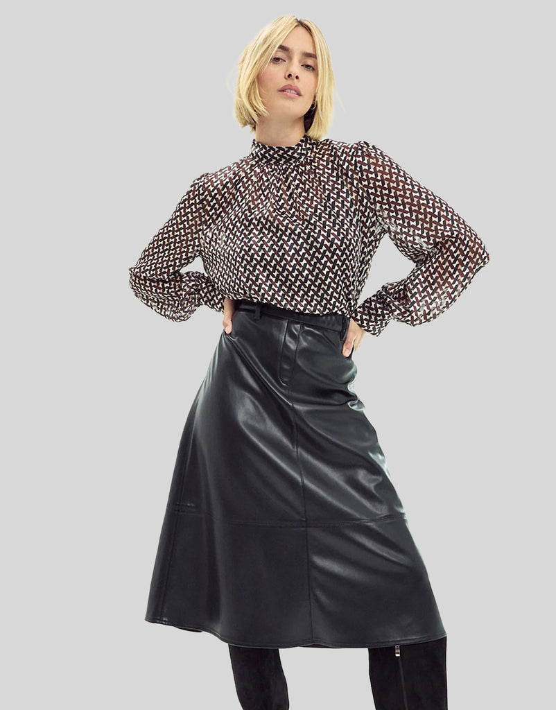 A model wearing a sheer black and white geometric patterned long-sleeve blouse with puffed shoulders, paired with a black faux-leather A-line skirt and black pants, set against a solid gray studio background.