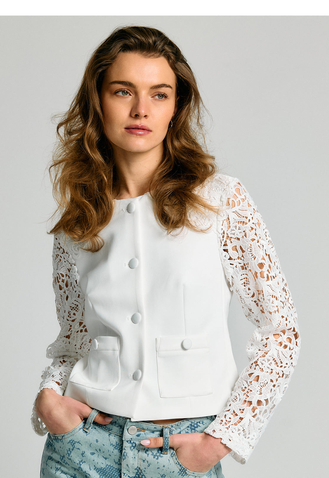 A woman modeling a white structured jacket with long, sheer floral lace sleeves, paired with light blue patterned jeans, against a solid gray studio background.