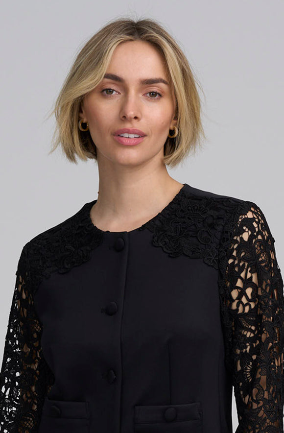 A woman with short blonde hair wearing a black long-sleeve top with lace yoke and lace sleeves, photographed against a plain light gray studio background.
