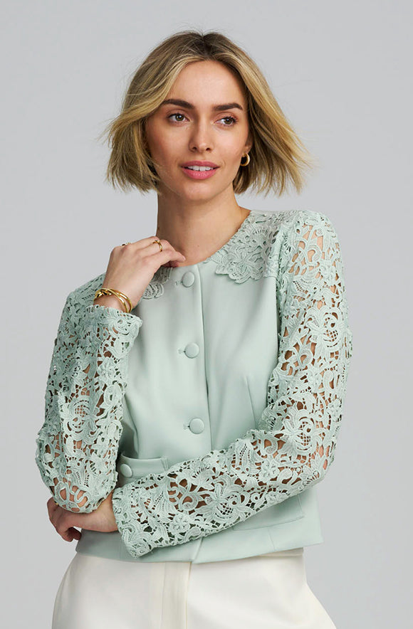 A woman modeling a mint green jacket with long lace sleeves and a button-front closure, paired with white bottoms, against a plain light gray studio background.