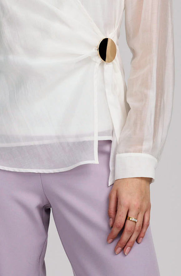 A close-up of a person wearing a white sheer long-sleeve blouse with a large round gold-tone button detail at the waist, paired with light lavender pants.